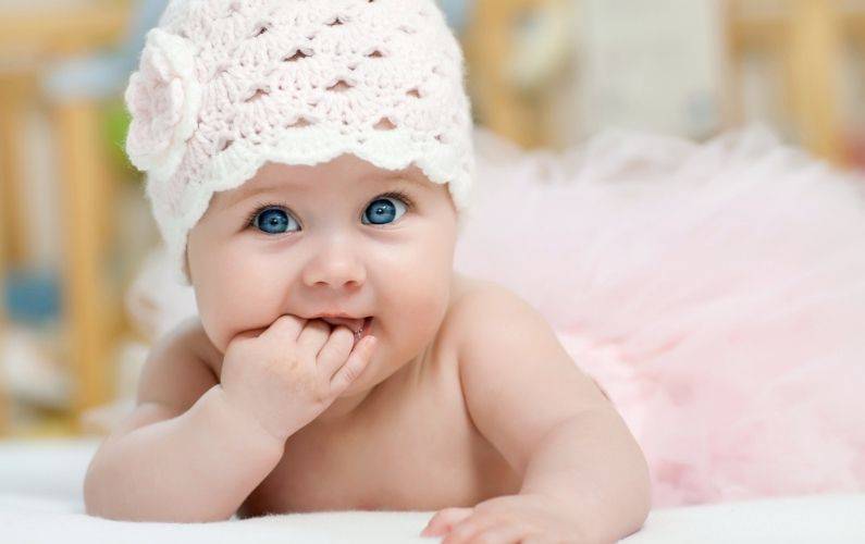 baby girl with hand in mouth while wearing a white knitted hat