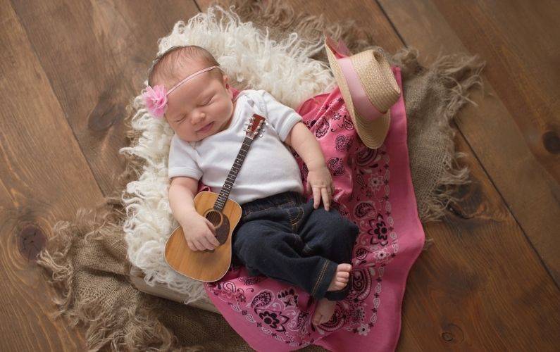 baby laying down with a pink bandana, guitar, and cowgirl hat